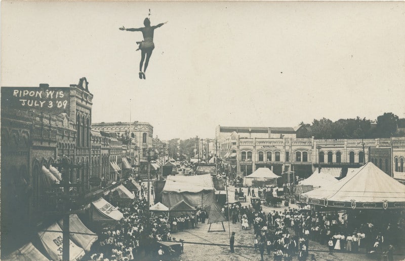 In this postcard, a daring acrobat dangles high above Ripon's central square. Based on the date (July 3), the tents, and the large crowd gathered below, this performance was most likely part of an Independence Day carnival in Ripon. Ripon Historical Society.