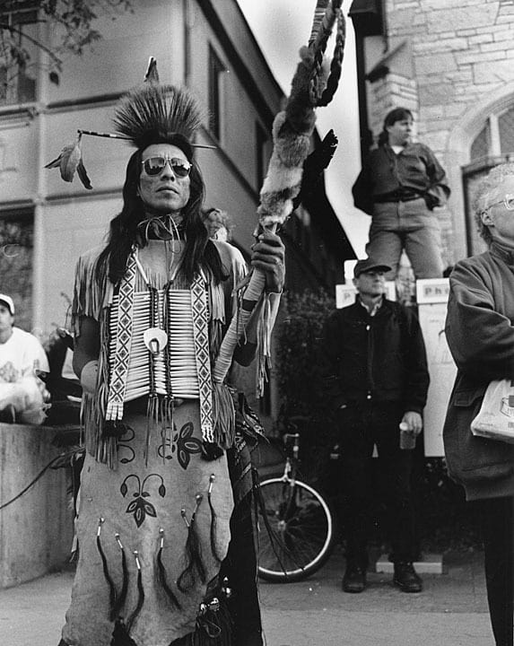 Art Shegonee, executive director of the Native American Center, watches speakers during a rally celebrating cultural resistance and survival on Columbus Day, 1992. UW-Madison Archives by way of University of Wisconsin Digital Collections.