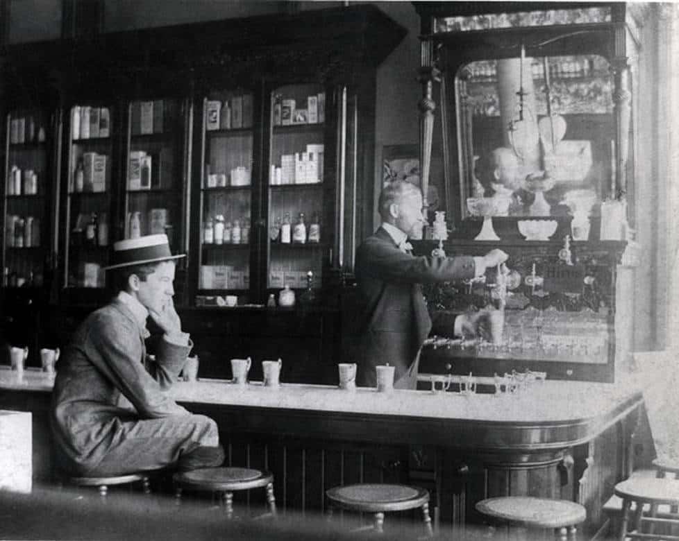 John Milton Granger sits on a caned stool at the marble-topped counter of Sykes Drug Store in Milwaukee, 1890s. Note the medicine bottles in the glass case next to the soda fountain. Milwaukee Public Library.