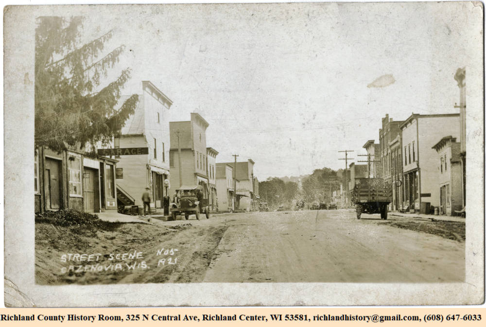 Black and white image looking down a street in Cazenovia with downtown buildings on both sides of street along with automobiles and utility poles. The car on the left side is being filled with gasoline. 1921.