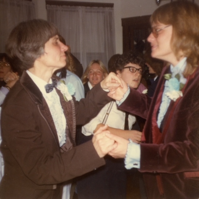 First Tux Party in 1980. Two women in brown tuxes are dancing together. In the background, more women can be seen dancing in the living room.