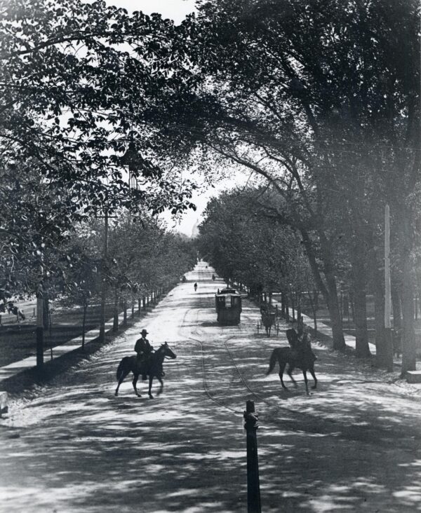 Two men ride horses across the streetcar tracks at the west end of State Street, a short distance from a streetcar. The street is tree-lined and the State Capitol is visible in the distance. May 1896.