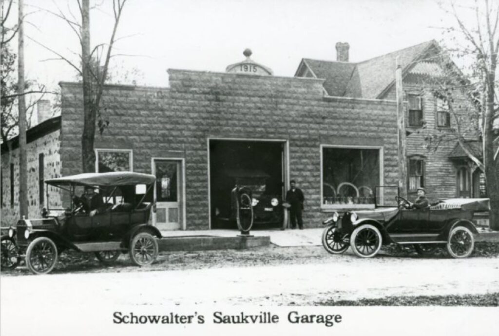 Image shows few vehicles at the Schowalter's Garage in Saukville, Wisconsin. The photo is dated as 1915. A handwritten note attached to the print indicates the house on the right to be Ben Mueller's. Photograph seems to be a copy from a postcard.