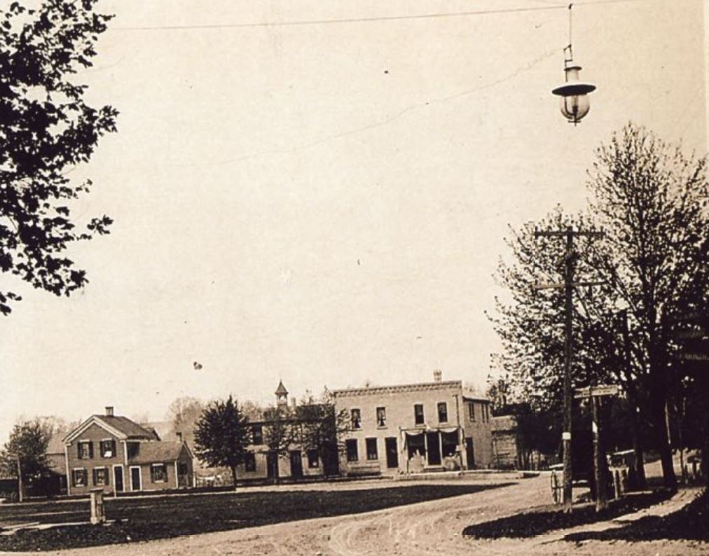 Image shows a section of Triangle Park, known too as Veterans Park in the foreground, with houses on the background which are facing South Mill street. A street lamp hanging from wires can be seen in the upper right corner of the photo. Undated.