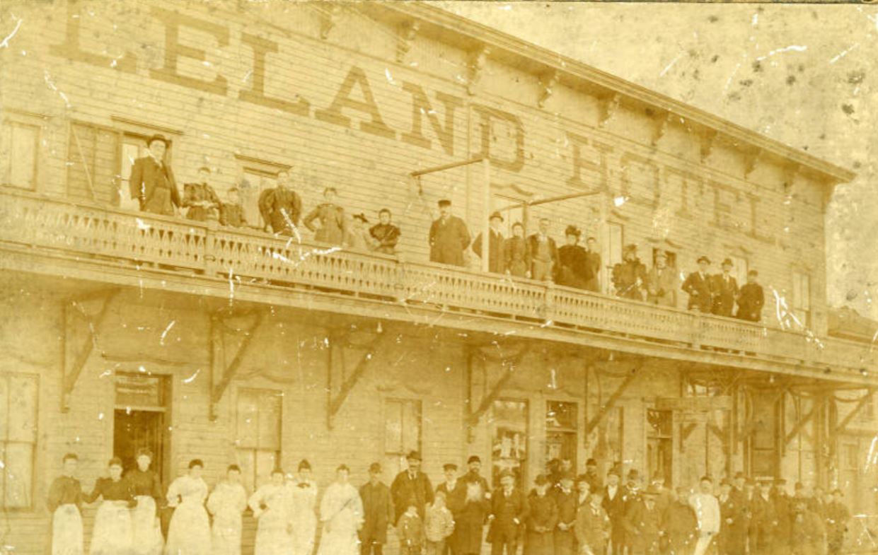 Exterior view of a wooden two story building with lettering at the top that says "Leland Hotel." Men, women and children stand out front and on the second story balcony.