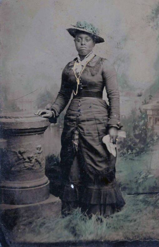 Portrait of unidentified woman wearing long dress and hat. Hand-tinting has been added to image.