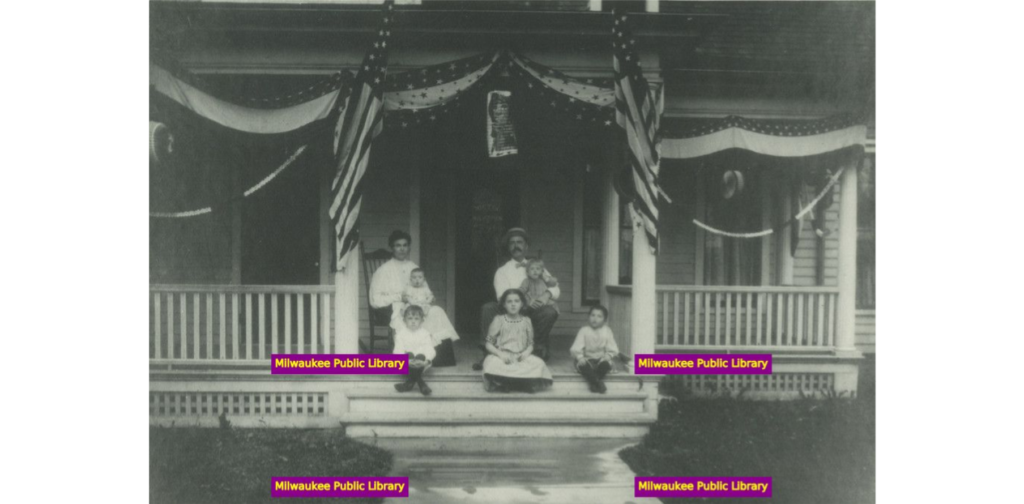 Family sitting on porch of house decorated for 4th of July, 1906-1907