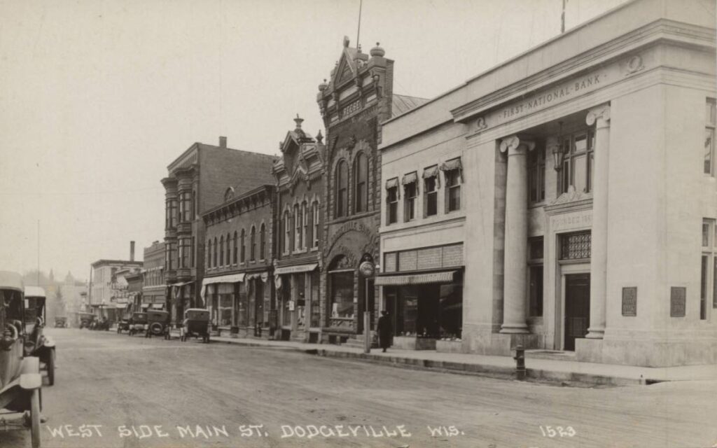 Iowa Street, including the First National Bank completed in 1919, and the old Dodgeville Bank, also known as the Reese Building. Past these are The Auditorium and Hotel Higbee as well as a drug store called Roberts and Prideaux. ca. 1925