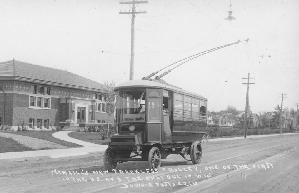 Merrill's trackless Trolley with T.B. Scott library in background, had solid rubber tires. It extended the street railway beyond the end of its rails to the sixth ward. Ca. 1900-1919.