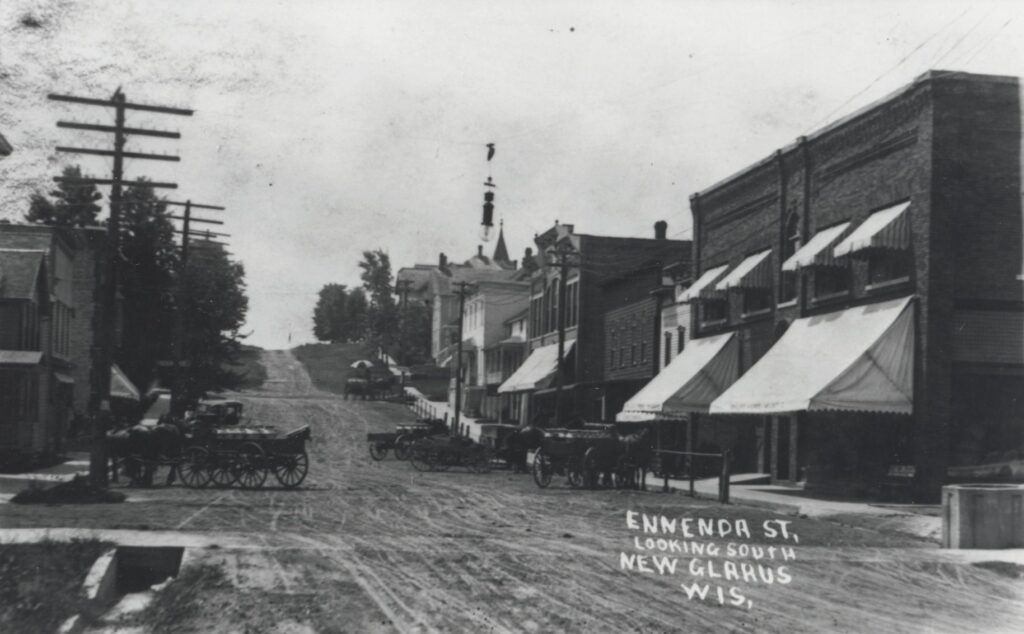 Horse-drawn wagons wait outside businesses on Second Street in New Glarus, then called Ennenda Street. The photo is looking south on Second Street from Fourth Avenue. ca. 1915
