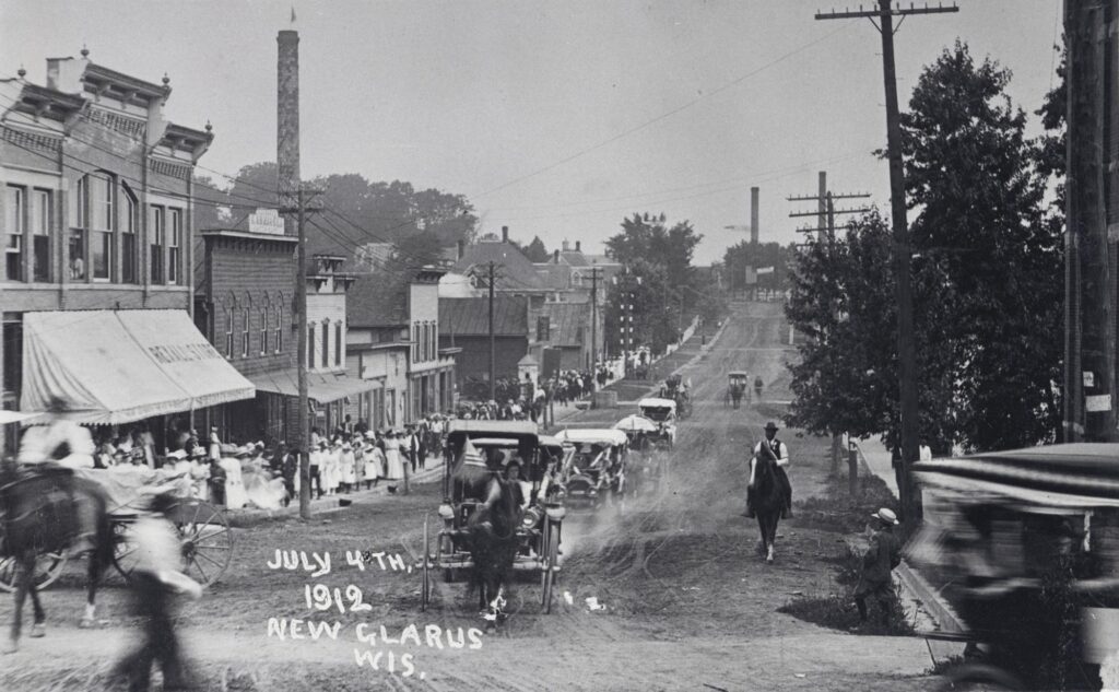 Onlookers watch a procession of automobiles and horse-drawn carriages during an Independence Day celebration in New Glarus, Wisconsin, on July 4, 1912. The photo was taken at the intersection of Second Street and Fifth Avenue, looking north on Second Street.