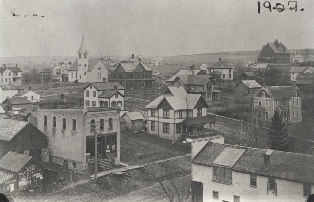 View of New Glarus looking southwest from Fifth Avenue near Second Street. The large brick building in the distance on the right is the New Glarus School. The white church on the left was first a Methodist church and then the Evangelical United Brethren Church. The building is now the Masonic Lodge. The house with a wraparound porch in the center of the photo now houses Deininger's Restaurant. The rectangular building on the left was the print shop of the New Glarus Post newspaper. 1907.