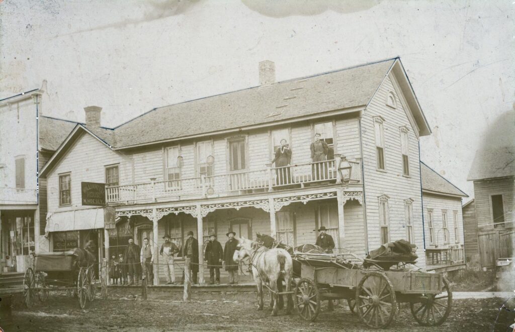 Men, women, and children stand on the porches and a horse-drawn wagon and a horse-drawn buggy stand in front of the William Tell House hotel before 1910. The hotel was located on Second Street between First Avenue and Second Avenue, where Flannery's Wilhelm Tell Restaurant is now located.