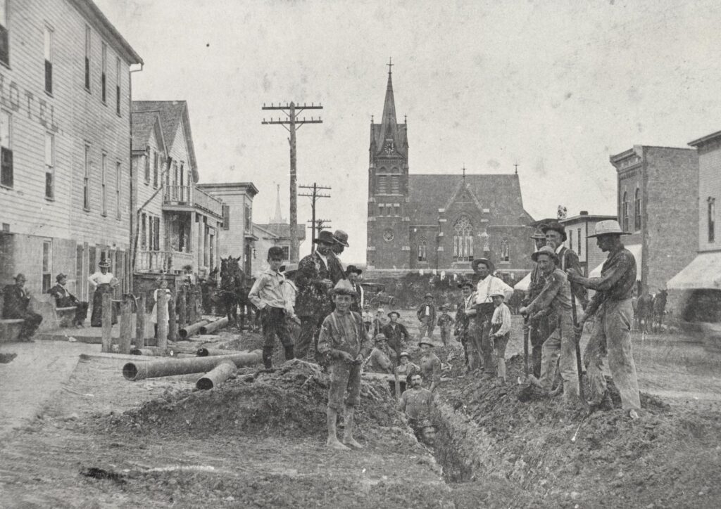 Workers excavate First Street in New Glarus, Wisconsin, to install water lines, as men, women, and children watch their progress. Horses and carriages can be seen in the background. The photo is looking north on First Street from the intersection with Sixth Avenue. Directly ahead is the brick Swiss Church, which was built in 1900, and the building on the left is the New Glarus Hotel. 1902.