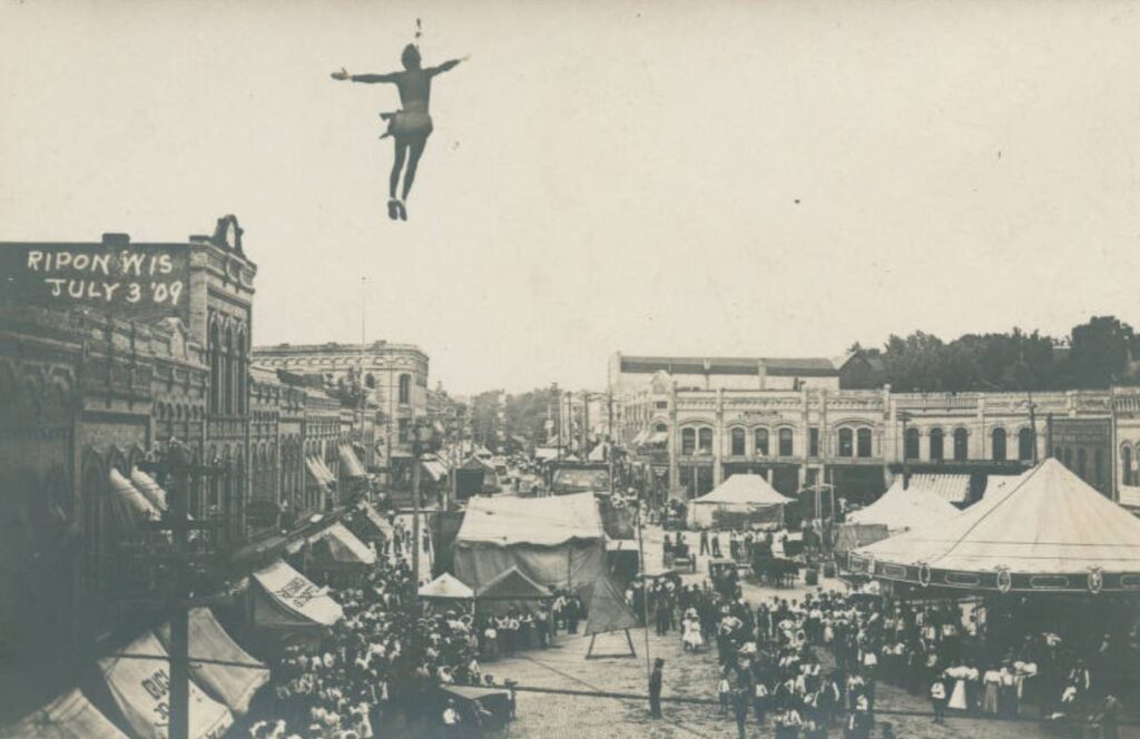 Carnival on Ripon's square with a trapeze artist hanging from her teeth from a cable, 1909.