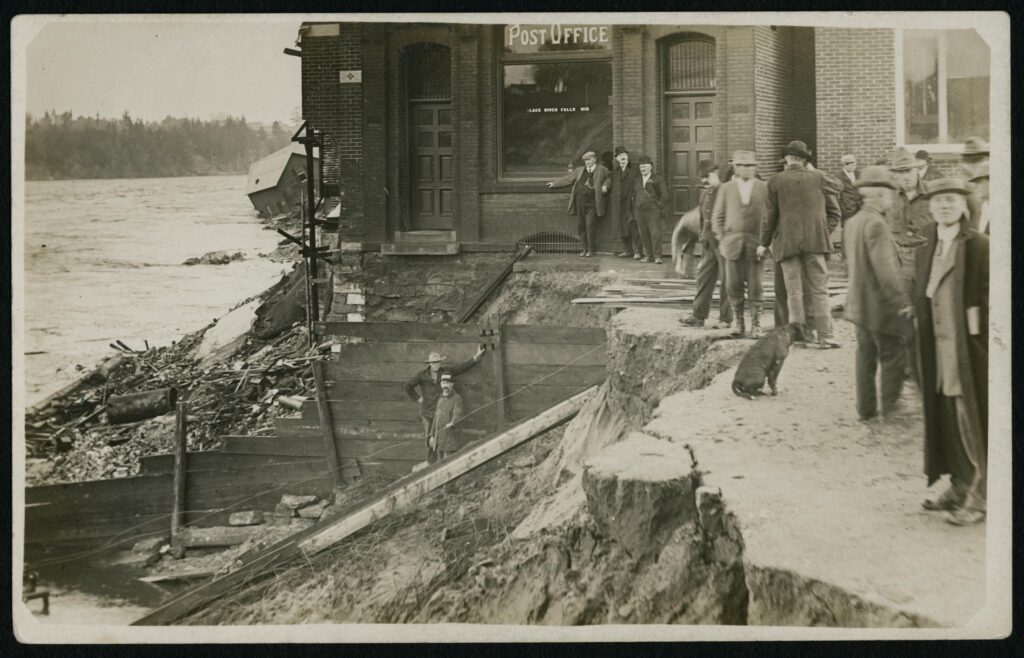 A postcard of a flood scene with a group of unidentified people and a dog gathered around what remains of the post office in Black River Falls, Wisconsin in 1911.