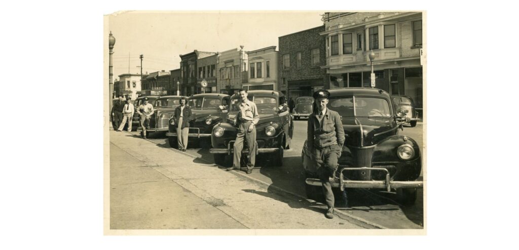 South Milwaukee Cab Company Fleet and Drivers, 1946. The fleet of the South Milwaukee Cab Company with drivers. The second driver from the right is identified as Chester Flancher. The Garden Theater is seen in the background.