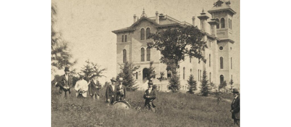 Exterior view of Old Main with a group of men with band instruments standing outside. Ca 1870.